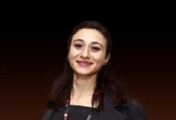 Portrait of a smiling young woman with dark hair wearing a black outfit against a dark gradient background.
