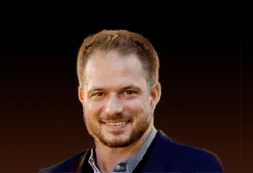 Portrait of a smiling man with short hair and a trimmed beard wearing a dark blazer against a dark gradient background.