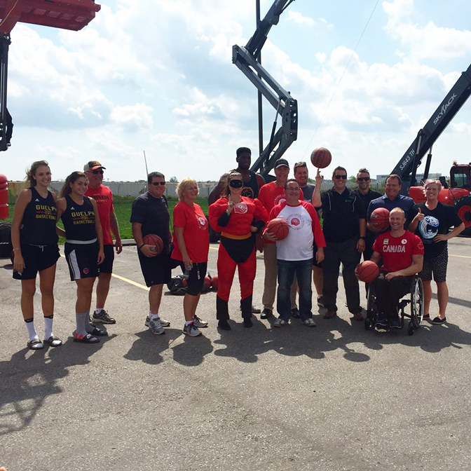 Group of people, including basketball players and supporters, posing outdoors near basketball hoop lifts on a sunny day.