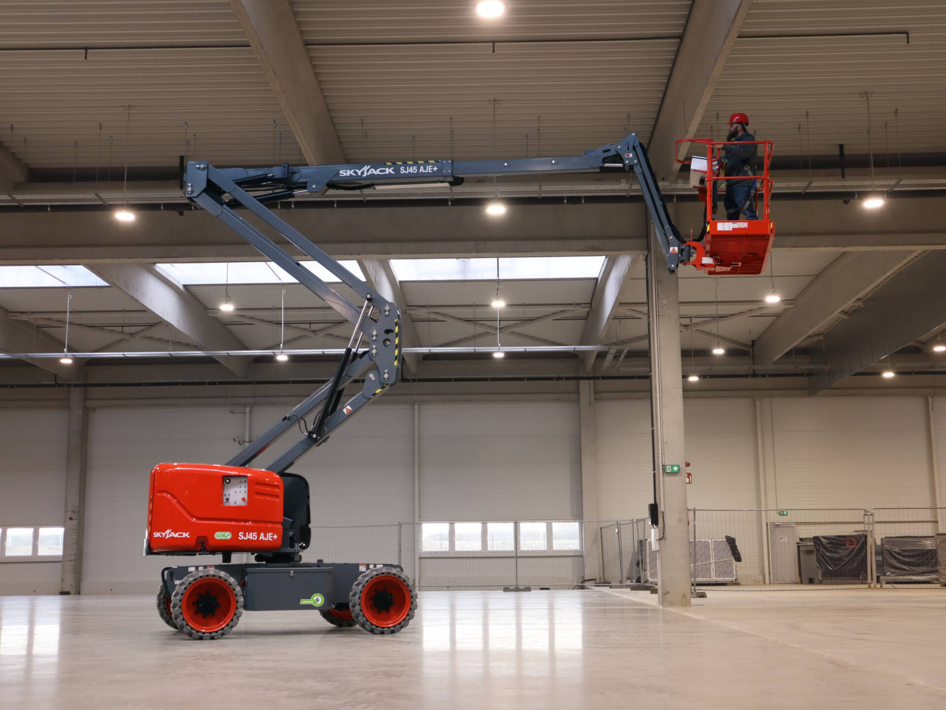 A red articulated boom lift with an extended arm and a worker in the basket operating inside a spacious industrial warehouse.