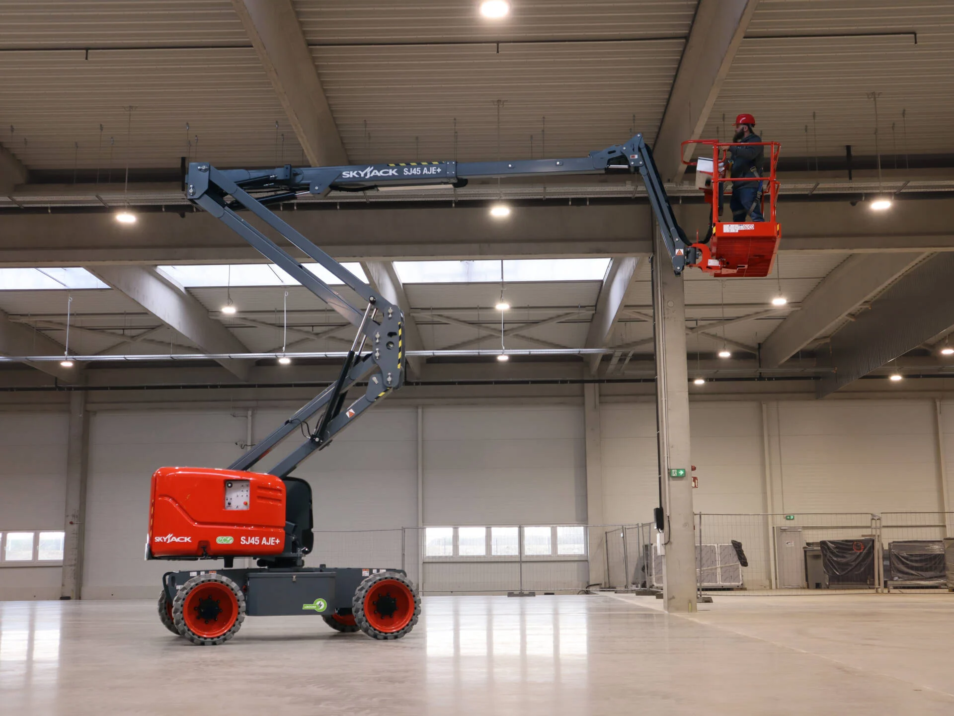 A red articulated boom lift with an extended arm and a worker in the basket operating inside a spacious industrial warehouse.