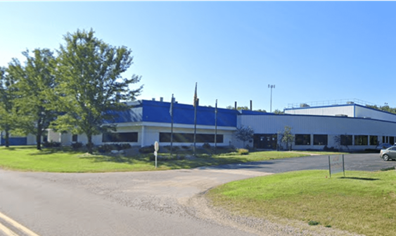 Exterior view of a modern industrial warehouse building with a blue and white facade, surrounded by trees and a grassy area under a clear sky.