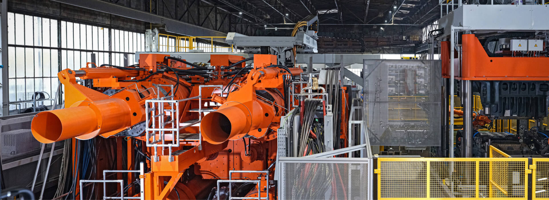Industrial orange robotic arms operate inside a spacious, modern manufacturing facility with large windows and metal structures.